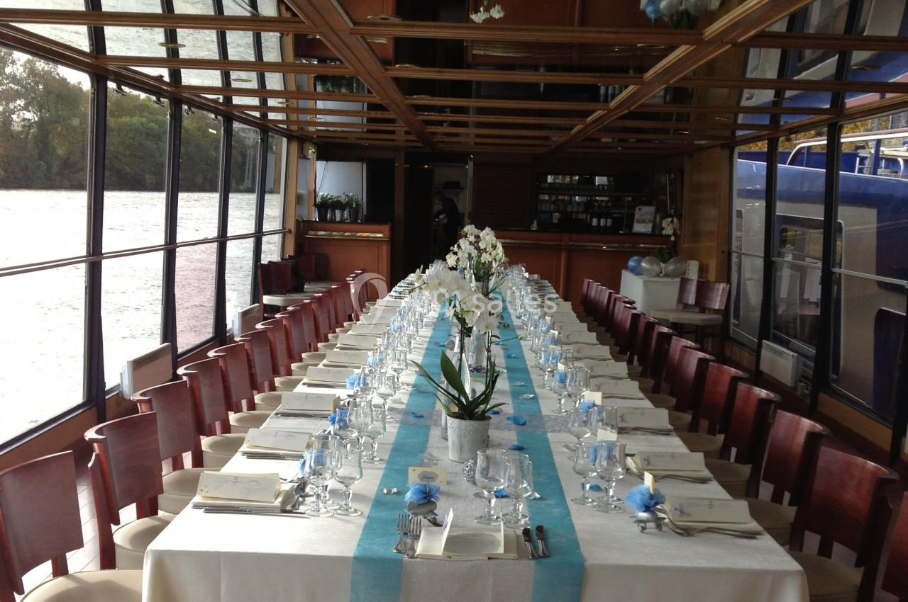 Salle de réception sur un bateau avec une longue table dressée, ornée de fleurs blanches et décorations bleues.
