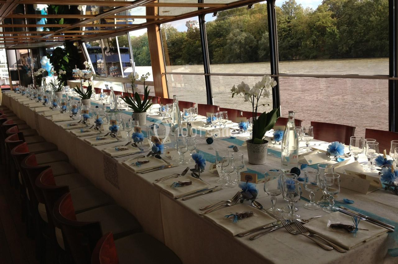 Table longue élégamment dressée avec des décorations bleues et blanches, située dans une salle avec vue sur une rivière.