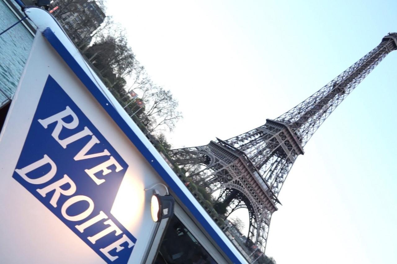 Bateau avec l'inscription ’Rive Droite’ au premier plan, et la tour Eiffel en arrière-plan sous un ciel clair.