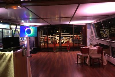Salle de restaurant sur un bateau avec tables rondes dressées, nappes blanches et vue sur un paysage fluvial.