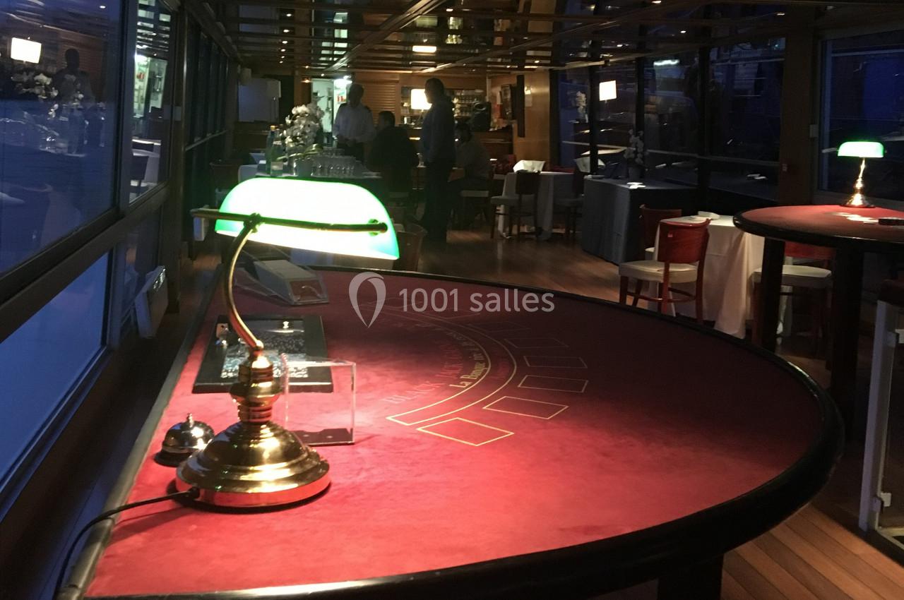 Table de jeu rouge éclairée par une lampe verte dans une salle avec des baies vitrées et un bar en arrière-plan.