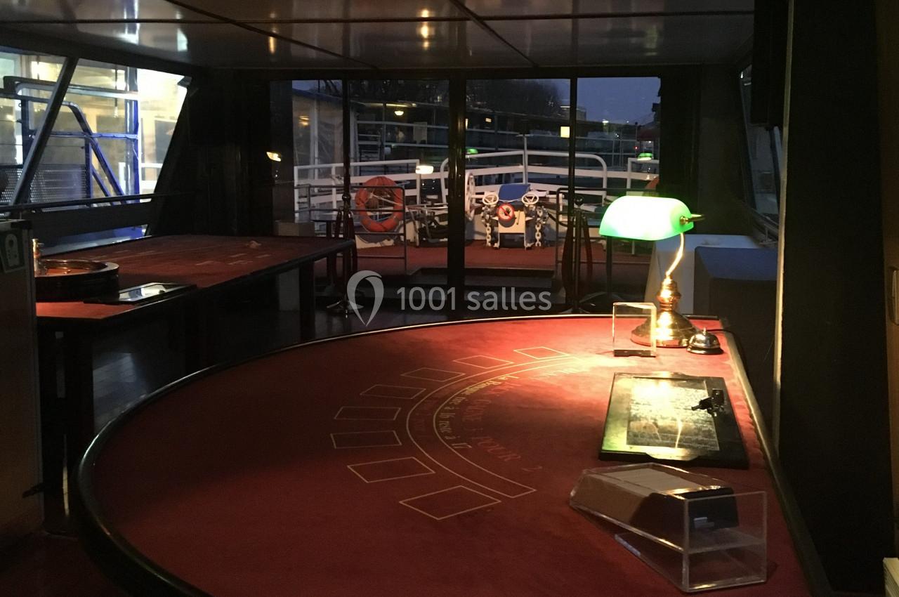Table de jeu de casino éclairée par une lampe, située dans une salle avec vue sur un pont extérieur.