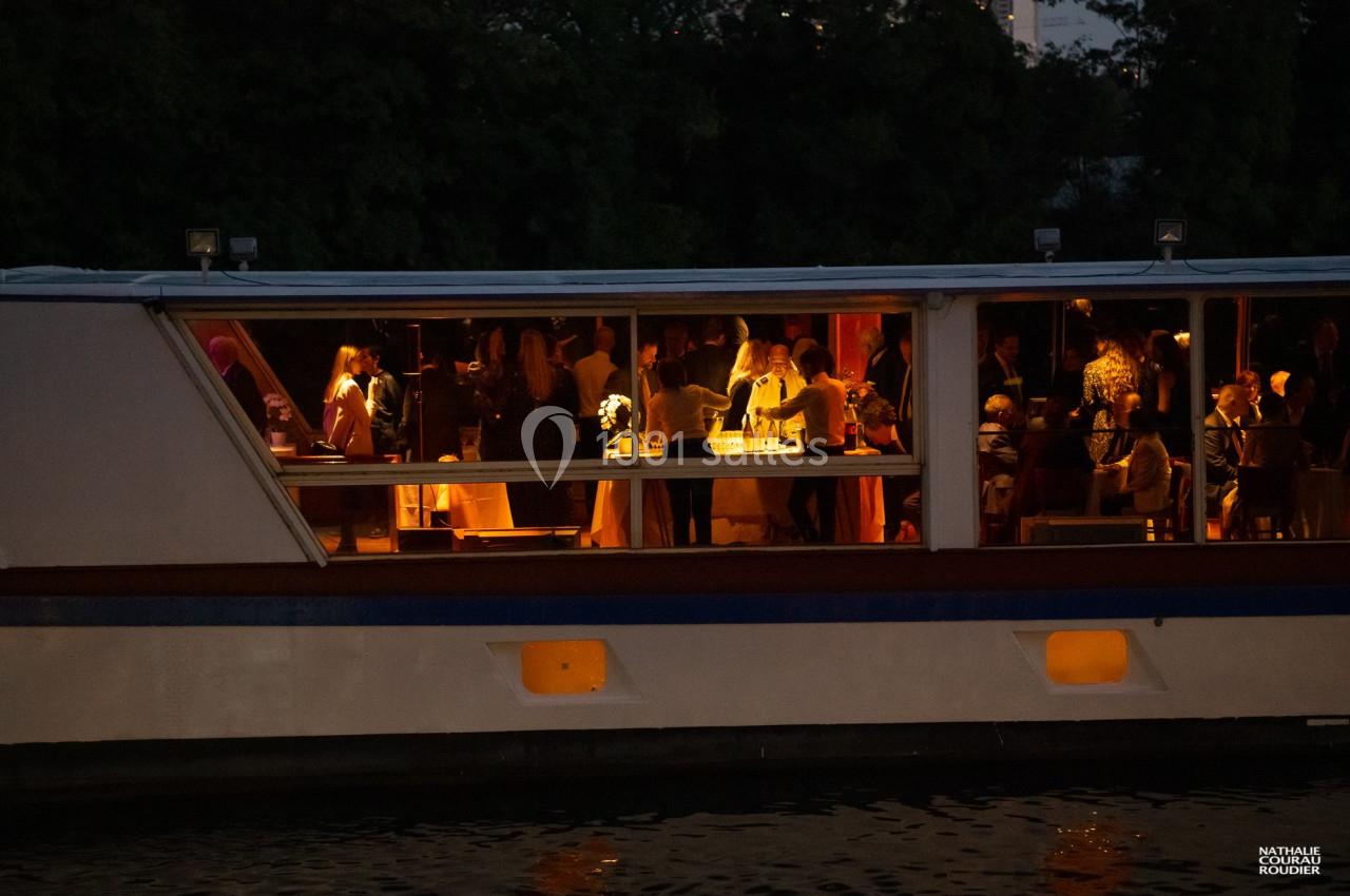 Des personnes participent à une réception éclairée à l'intérieur d'un bateau amarré, en soirée.