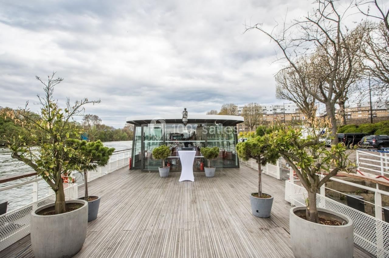 Terrasse en bois d'un bateau avec des plantes en pots, vue sur une salle vitrée et un fleuve en arrière-plan.