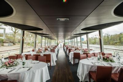 Salle de restaurant sur un bateau avec tables rondes dressées, nappes blanches et vue sur un paysage fluvial.