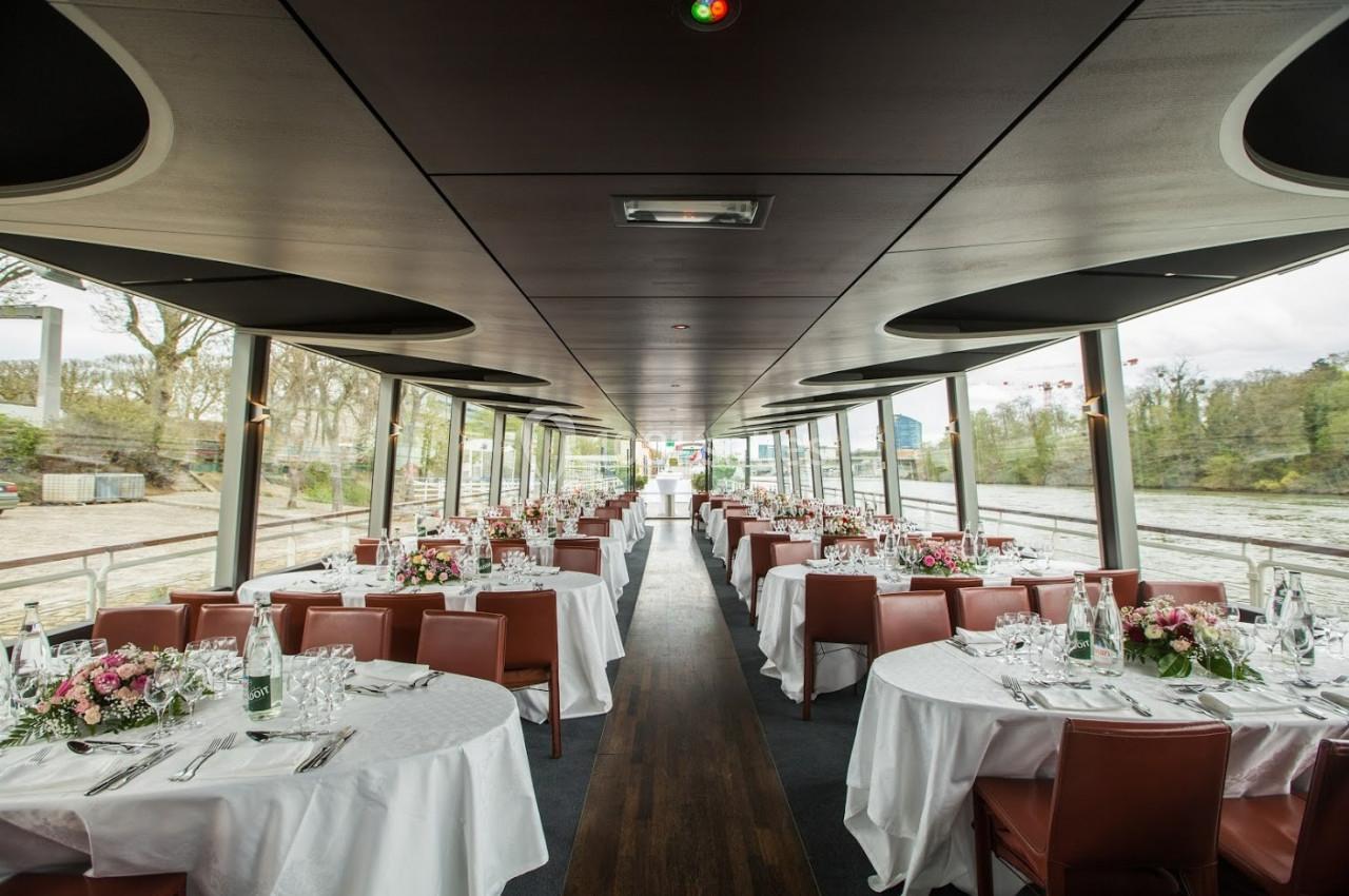 Salle de réception sur une péniche avec tables rondes dressées, nappes blanches et vue sur un paysage fluvial.