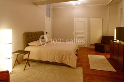 Lit double avec draps blancs dans une chambre mansardée, parquet au sol et poutres apparentes.