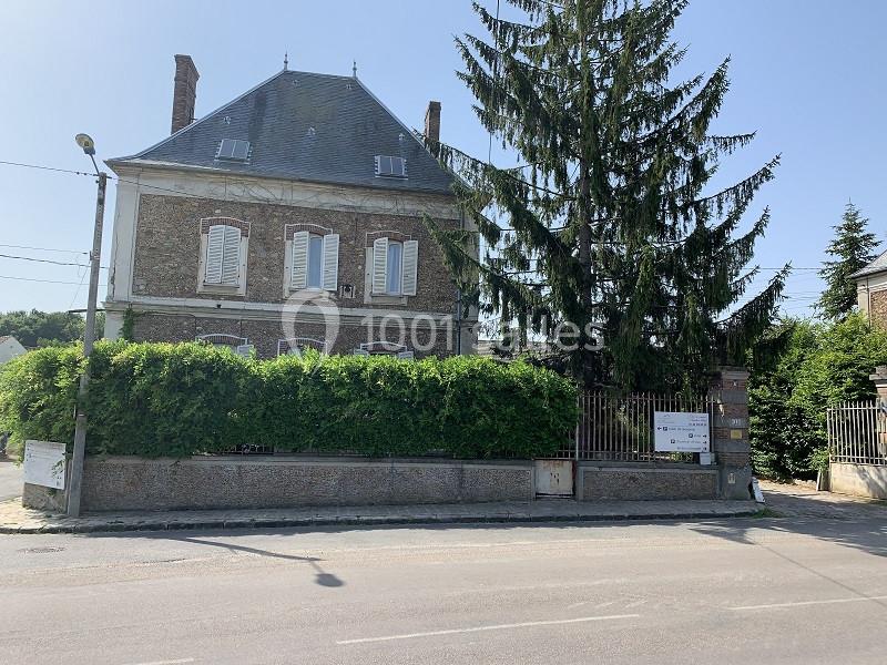 Façade d'une maison en briques avec volets blancs, entourée d'arbres et d'une haie, vue depuis une rue ensoleillée.