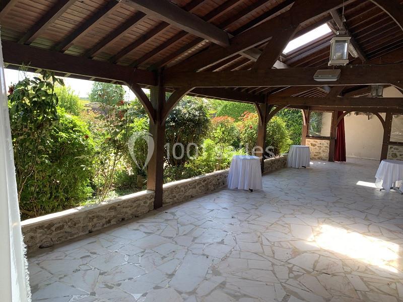 Terrasse couverte avec charpente en bois, sol en pierre et vue sur un jardin verdoyant.