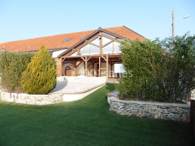 Façade d'un bâtiment avec toit en tuiles, grande verrière en bois, entouré de haies et d'une pelouse artificielle.