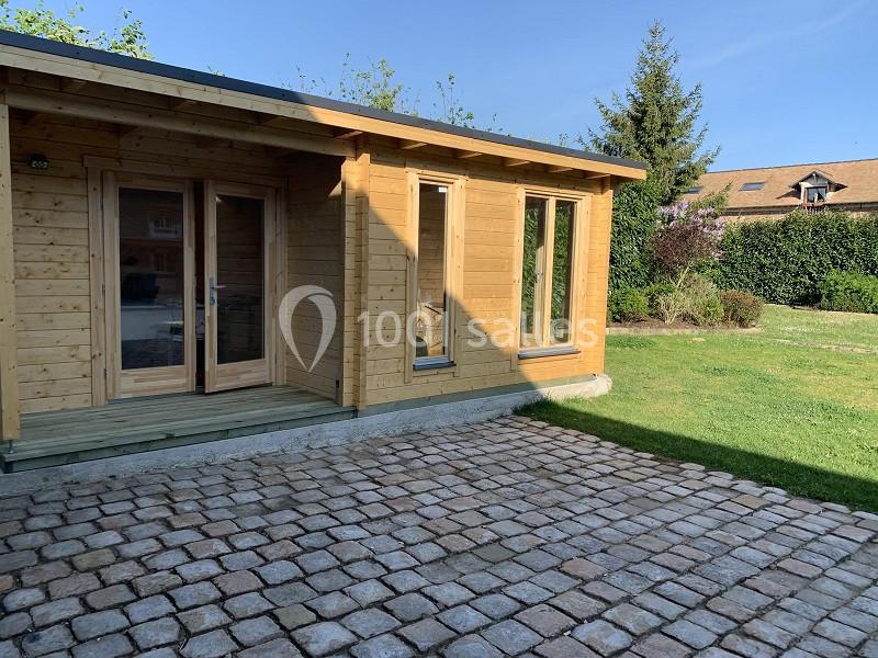 Chalet en bois avec terrasse pavée, entouré de verdure et d'arbres sous un ciel dégagé.