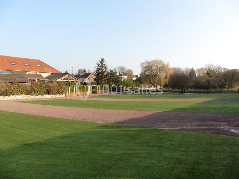 Terrain de sport en plein air avec pelouse et piste en terre, entouré de maisons et d'arbres sous un ciel dégagé.
