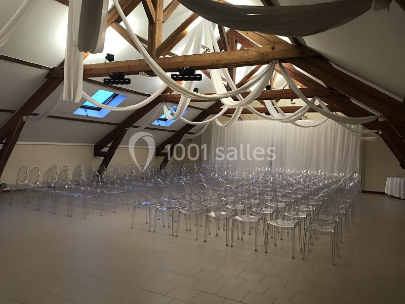 Salle de réception avec poutres apparentes, chaises transparentes alignées et drapés blancs au plafond.
