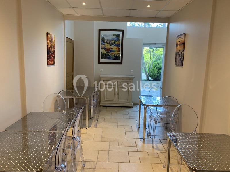 Salle lumineuse avec tables en métal, chaises transparentes, tableaux colorés aux murs et vue sur un jardin verdoyant.