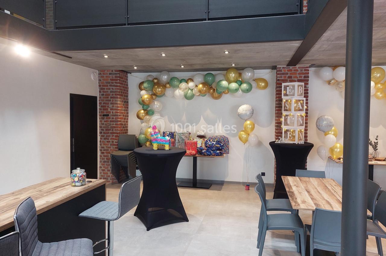 Salle décorée pour une fête avec ballons colorés, tables en bois, chaises grises et cadeaux posés sur une table.
