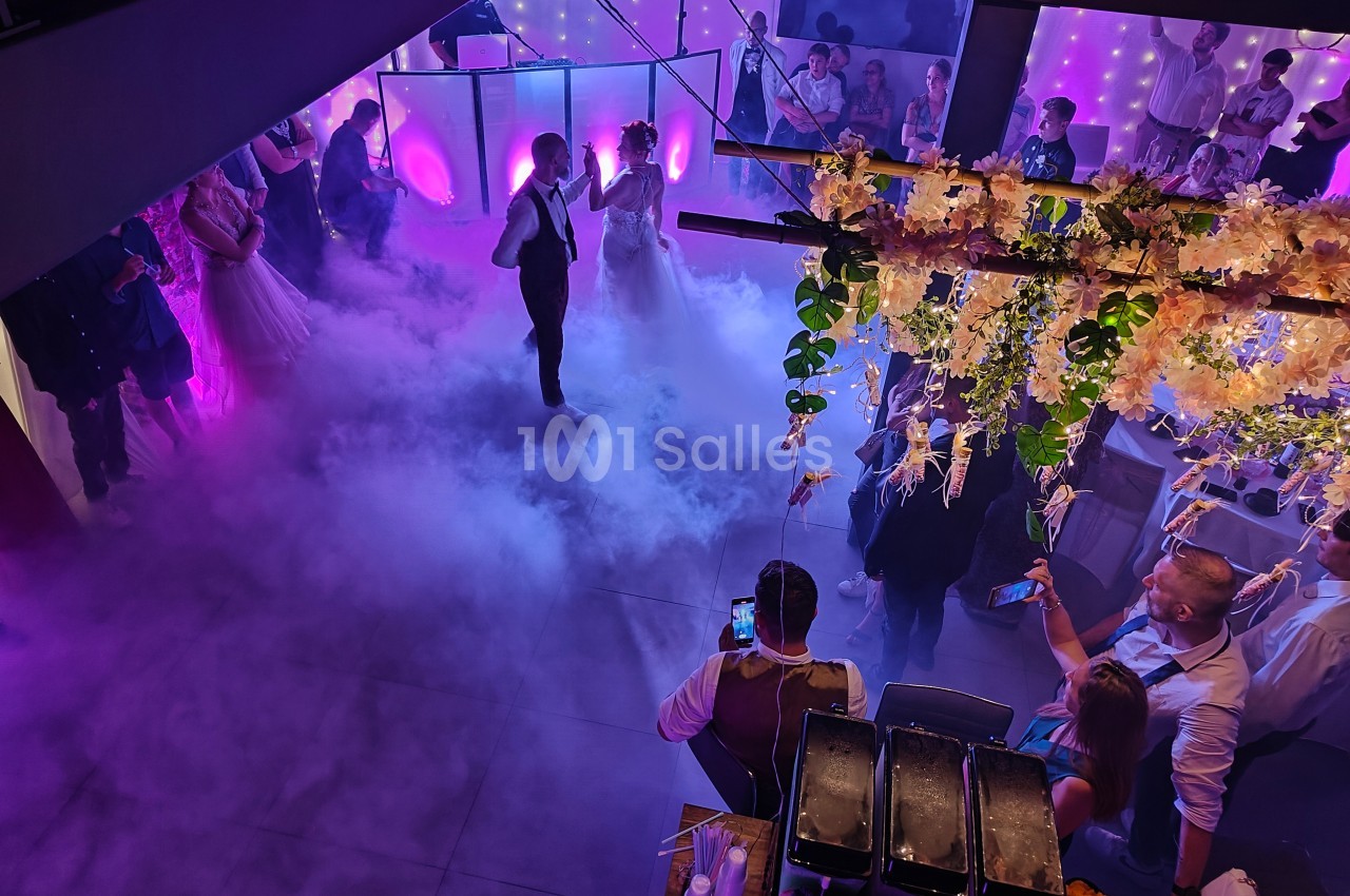 Un couple danse au centre d'une piste enveloppée de fumée, entouré d'invités et d'une lumière violette.