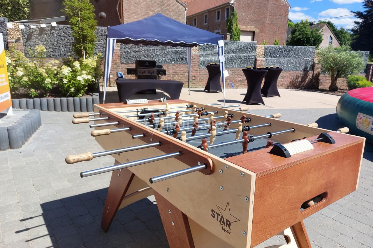 Table de baby-foot en bois installée en extérieur sur une terrasse pavée, avec un chapiteau et des tables en arrière-plan.