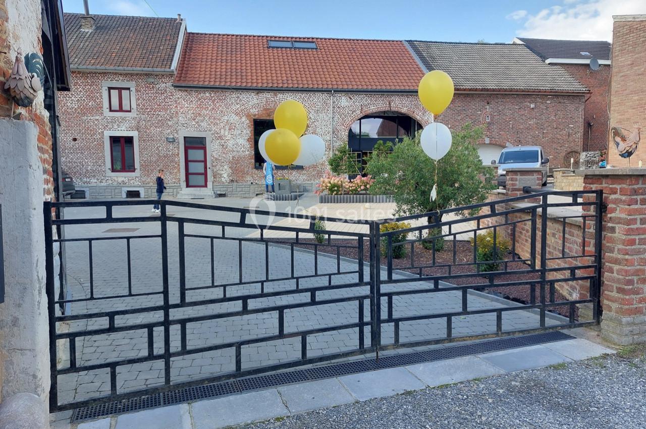 Portail métallique noir ouvert devant une cour pavée avec des ballons jaunes et blancs attachés à une clôture.