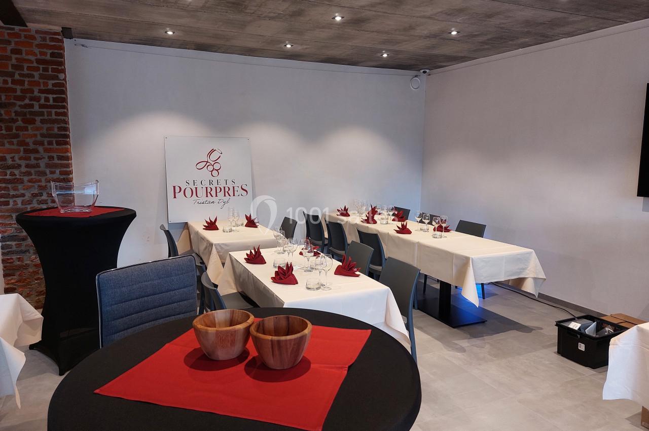 Salle de réception aménagée avec des tables dressées, nappes blanches, serviettes rouges et décoration sobre.