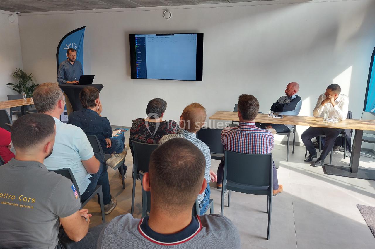 Un homme présente un contenu sur un écran à un groupe de personnes assises dans une salle lumineuse.