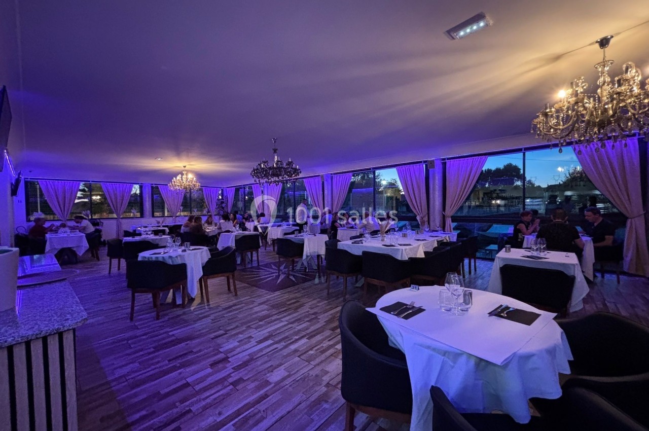 Salle de restaurant élégante avec éclairage tamisé violet, tables dressées et grandes baies vitrées donnant sur l'extérieur.