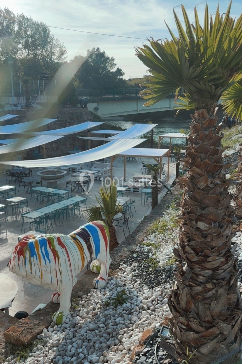 Statue de taureau colorée près d'une terrasse extérieure avec tables, chaises et palmiers, au bord d'une rivière.