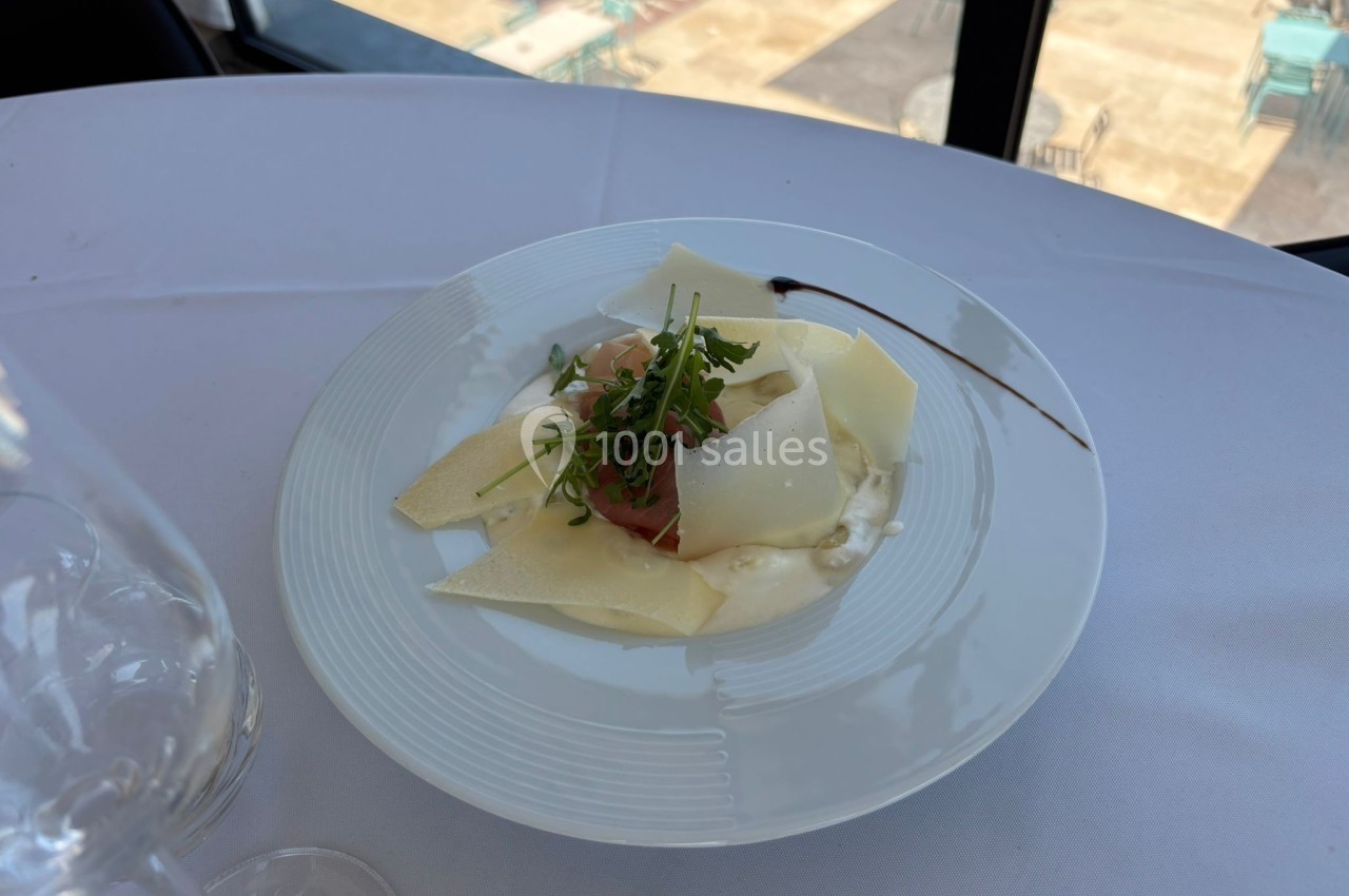 Assiette de carpaccio avec copeaux de parmesan, roquette et filet de sauce, servie sur une table blanche près d'une fenêtre.