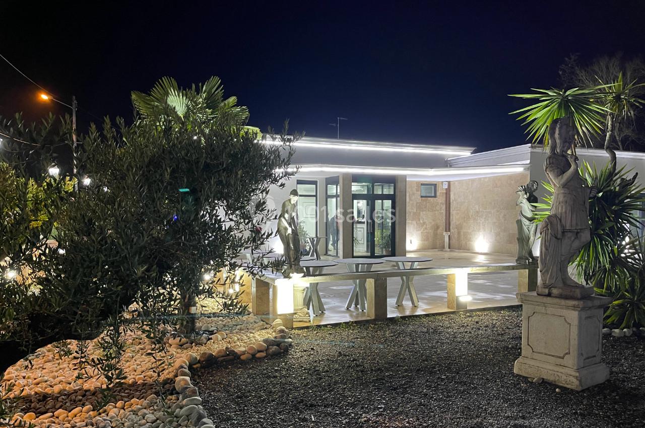 Entrée d'une maison moderne éclairée la nuit, entourée de statues, plantes et d'un jardin de galets.