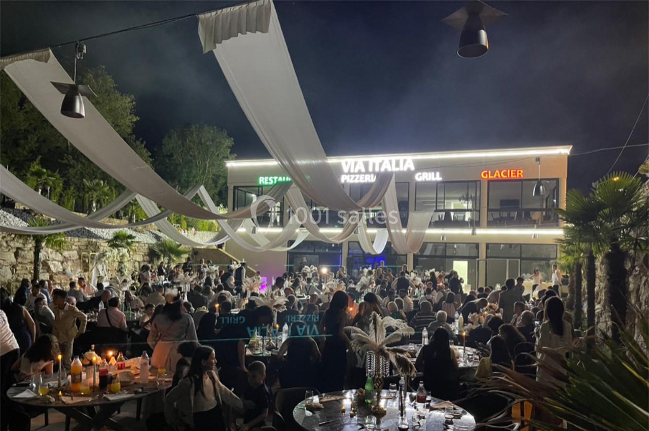 Foule rassemblée en soirée dans une terrasse éclairée, décorée de voilages blancs, devant un restaurant.