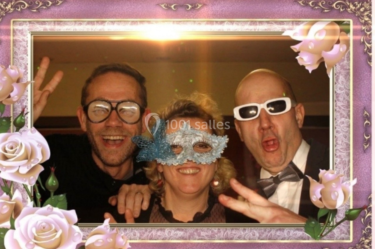 Trois personnes posent en souriant avec des accessoires festifs, encadrées par un décor rose orné de roses.