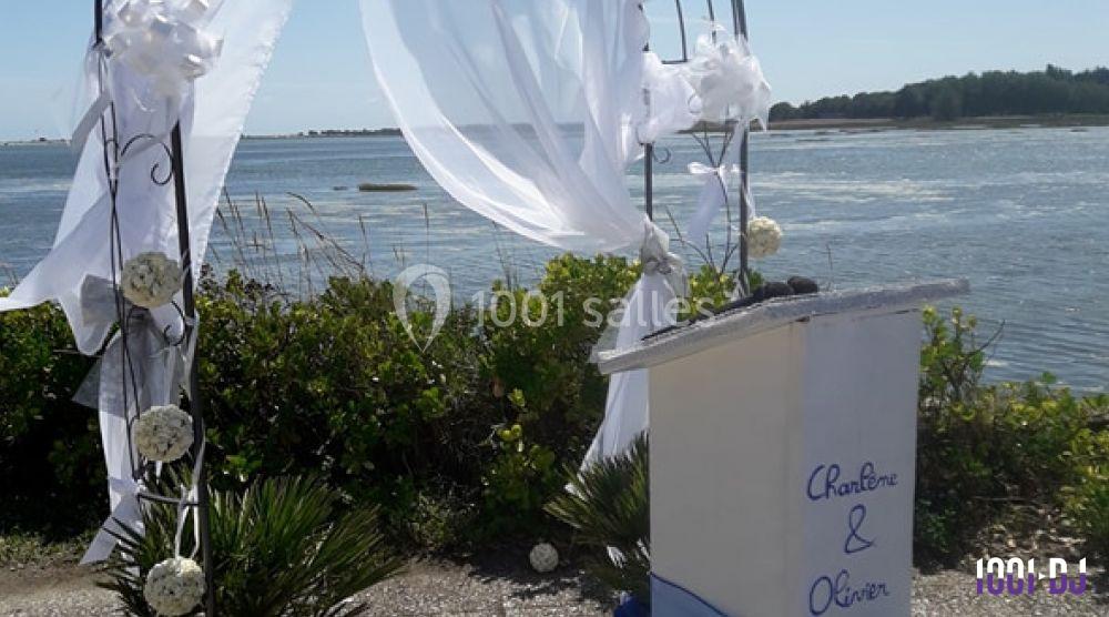 Arche décorée de voiles blancs et fleurs blanches, installée en extérieur près d'un plan d'eau.
