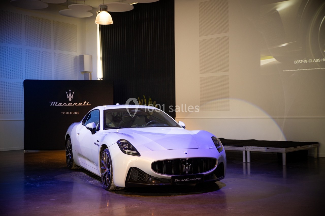 Voiture de sport blanche exposée dans une salle avec éclairage tamisé et panneau Maserati en arrière-plan.