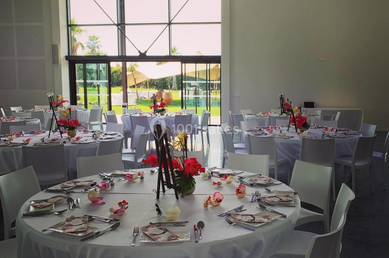 Salle lumineuse avec tables rondes dressées, nappes blanches, décorations florales et vue sur un espace extérieur verdoyant.