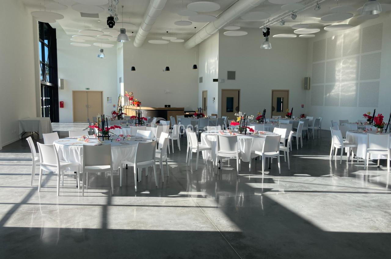 Salle lumineuse avec tables rondes dressées, chaises blanches et décorations florales colorées, prête pour un événement.