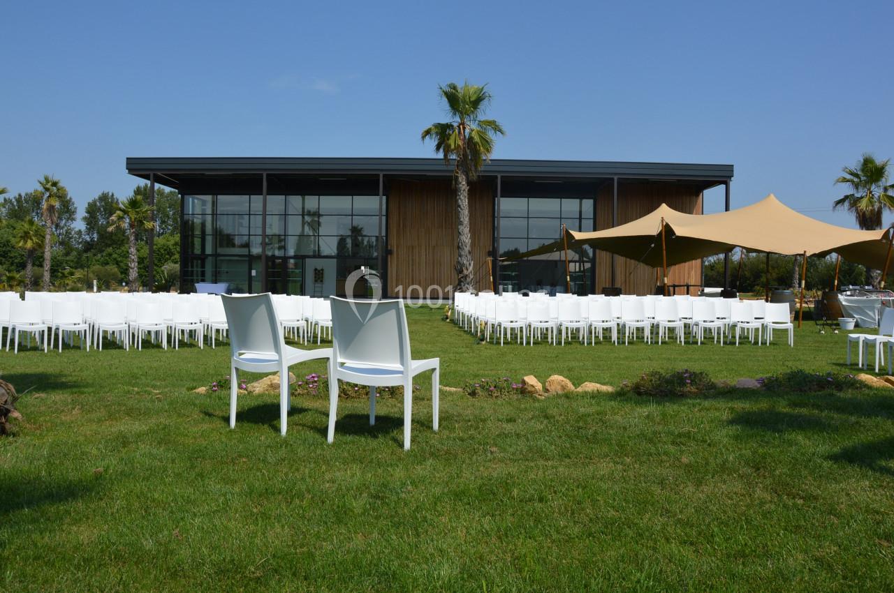 Façade moderne d'un bâtiment vitré entouré de chaises blanches disposées sur une pelouse avec des palmiers.