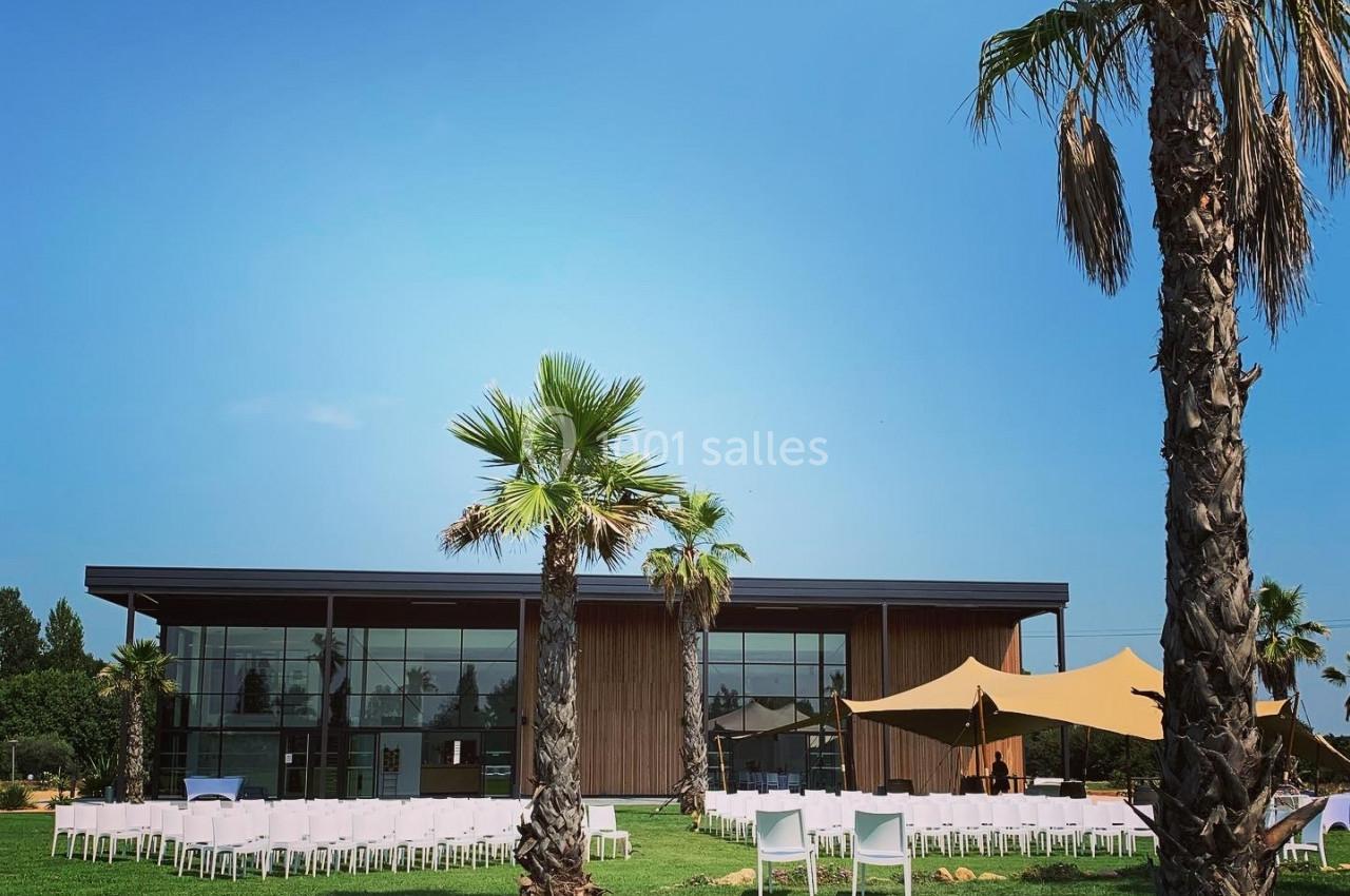 Extérieur d'un bâtiment moderne avec façade vitrée, entouré de palmiers et de chaises blanches sur une pelouse.