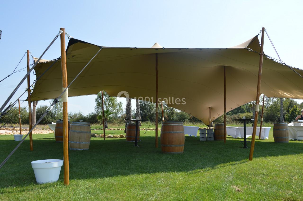 Tente beige tendue sur une pelouse, entourée de tonneaux en bois et de tables hautes, en extérieur.