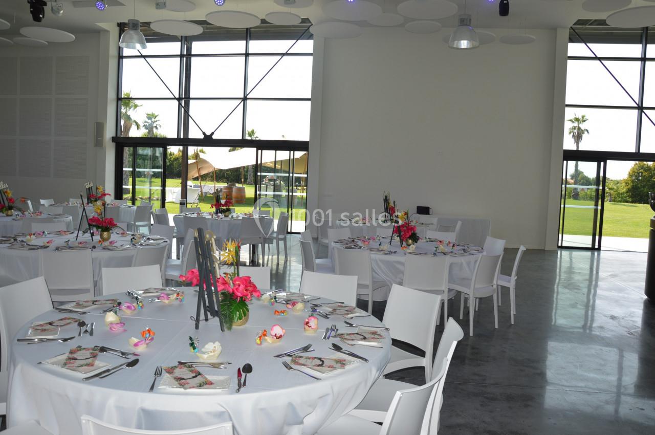 Salle lumineuse avec tables rondes dressées pour un événement, décorées de fleurs colorées, vue sur un jardin extérieur.