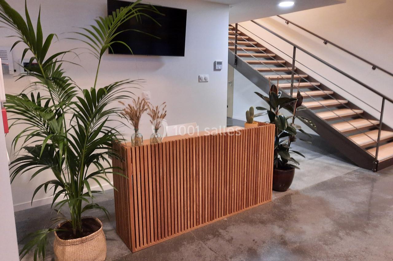 Accueil avec un comptoir en bois, plantes décoratives, escalier en arrière-plan et écran mural.