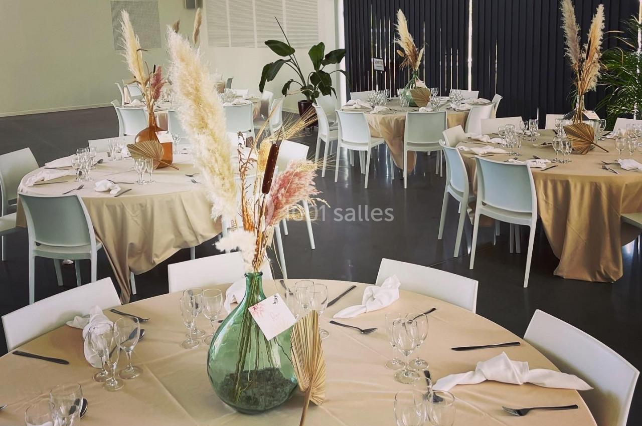 Salle de réception avec tables rondes dressées, nappes beige, chaises blanches et décorations florales élégantes.