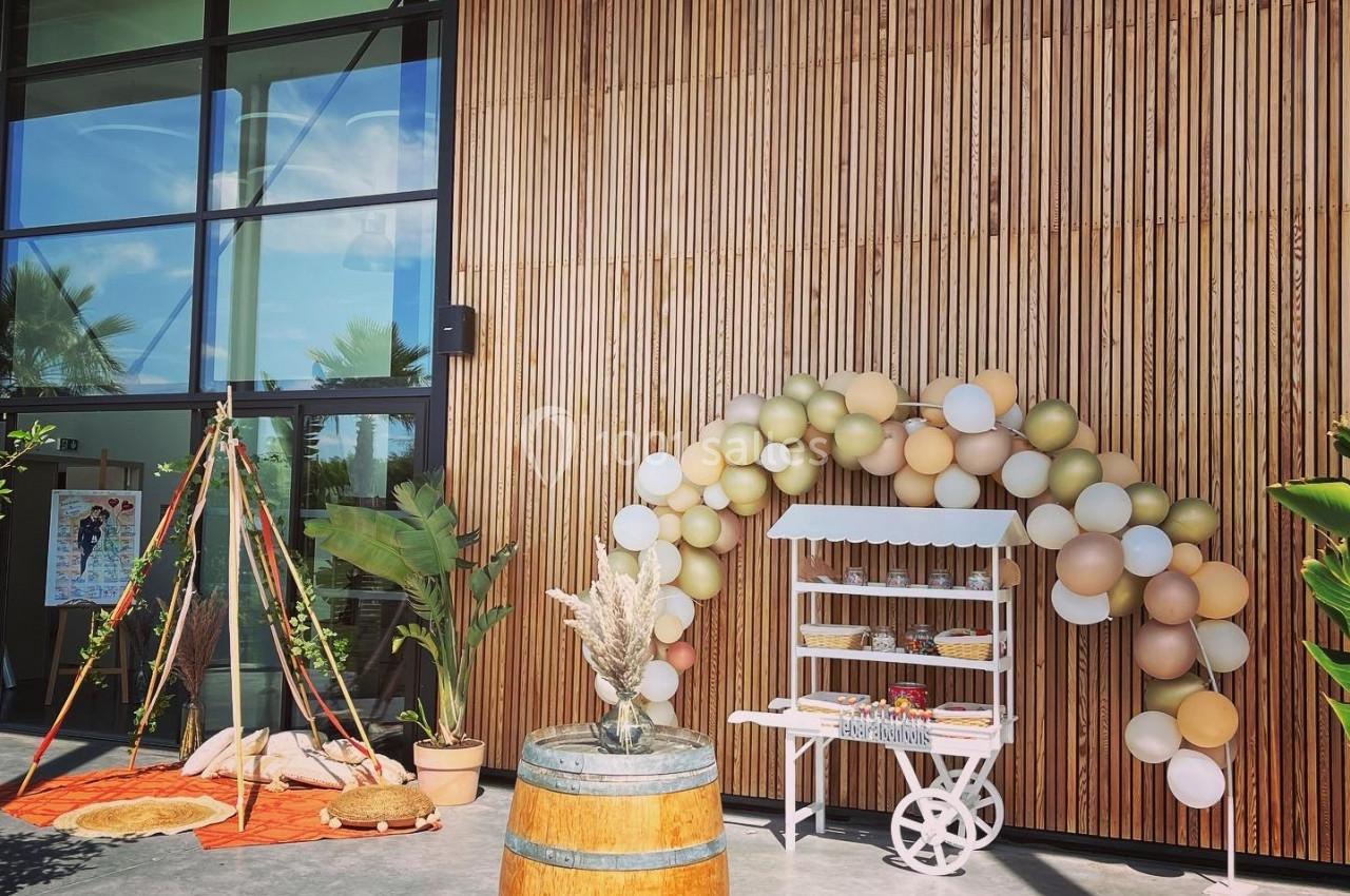 Décoration extérieure avec arche de ballons, tipi, tonneau en bois et étagère sur fond de façade en bois et verre.