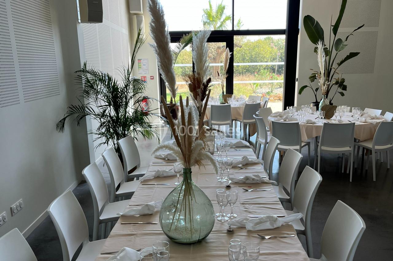 Salle lumineuse avec tables dressées, décorées de vases contenant des herbes séchées, et grandes baies vitrées donnant sur l…