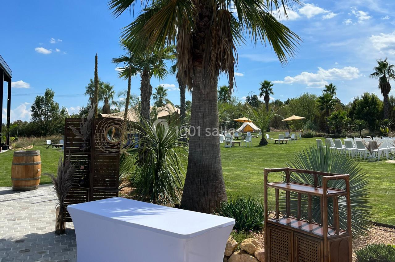 Jardin extérieur avec palmiers, pelouse, mobilier de jardin et table blanche sous un ciel ensoleillé.