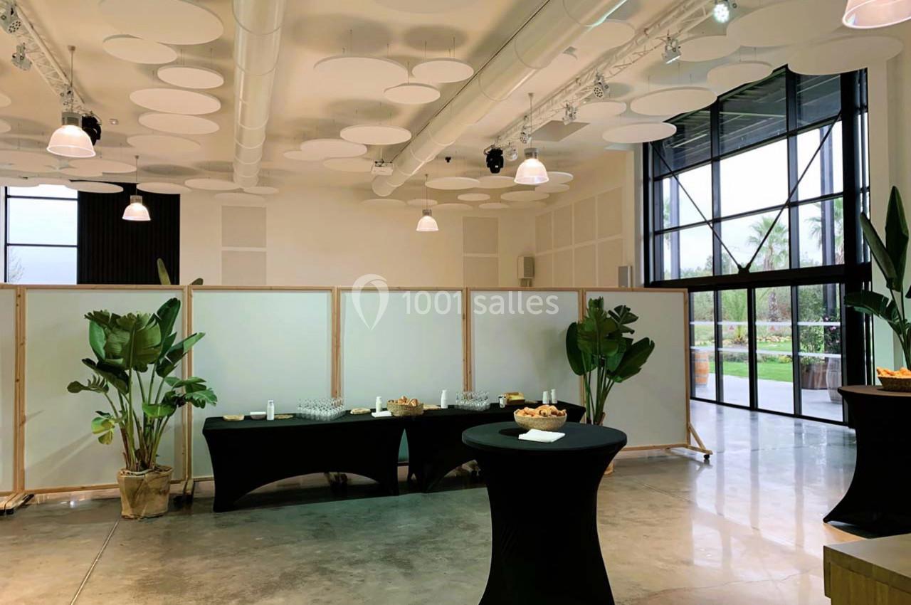Salle lumineuse avec tables hautes noires, plantes décoratives et buffet installé près de cloisons blanches.
