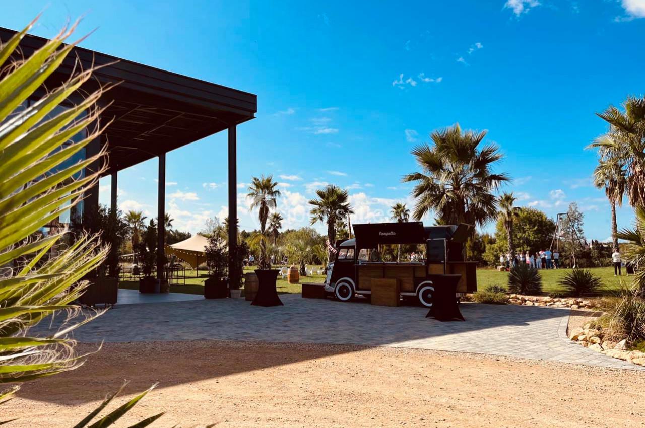 Un food truck noir stationné près d'une terrasse couverte, entouré de palmiers sous un ciel bleu.
