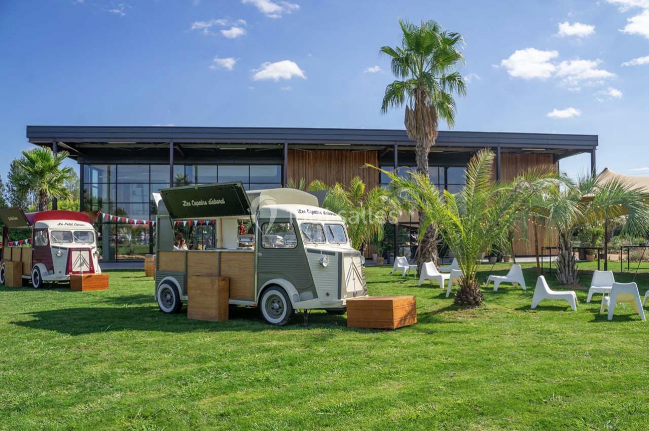 Deux food trucks vintage stationnés sur une pelouse devant un bâtiment moderne, entourés de palmiers et de chaises.