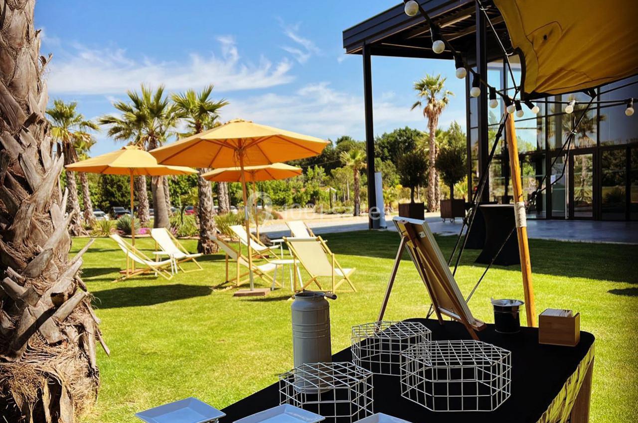 Table dressée en extérieur avec des assiettes et des décorations, vue sur un jardin avec transats et parasols jaunes.