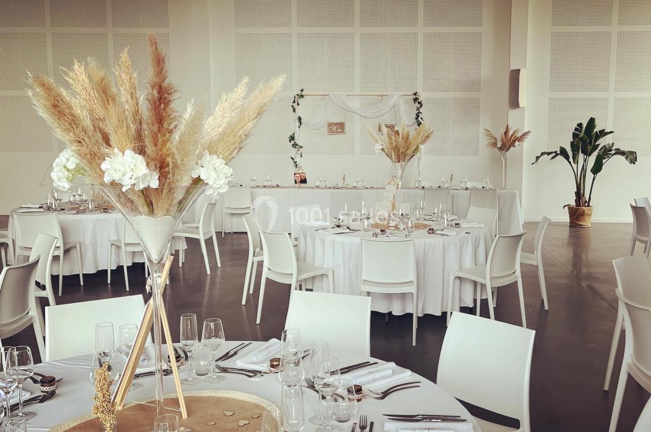 Salle de réception décorée pour un mariage, avec tables rondes dressées et ornées de bouquets de fleurs séchées.