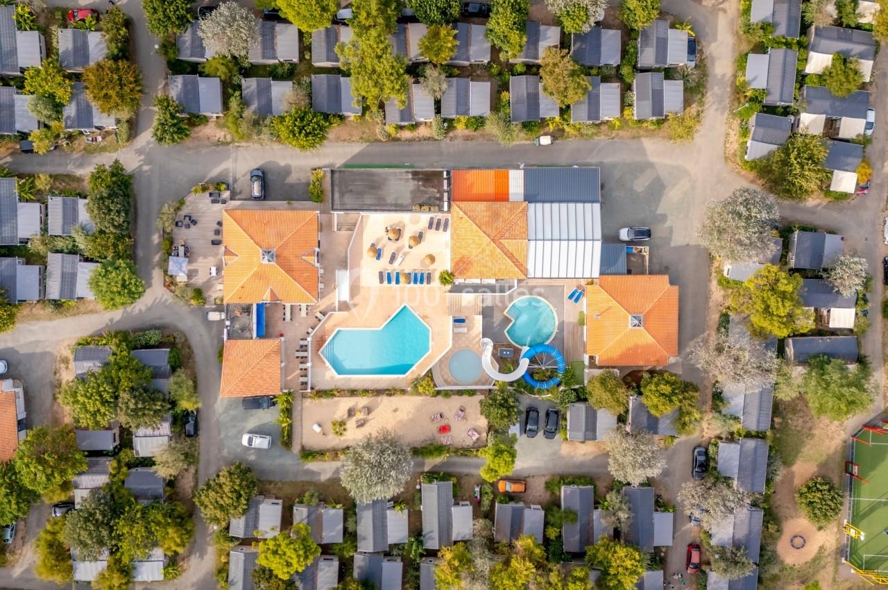 Vue aérienne d'un complexe avec piscine, toits en tuiles et mobil-homes entourés de végétation.