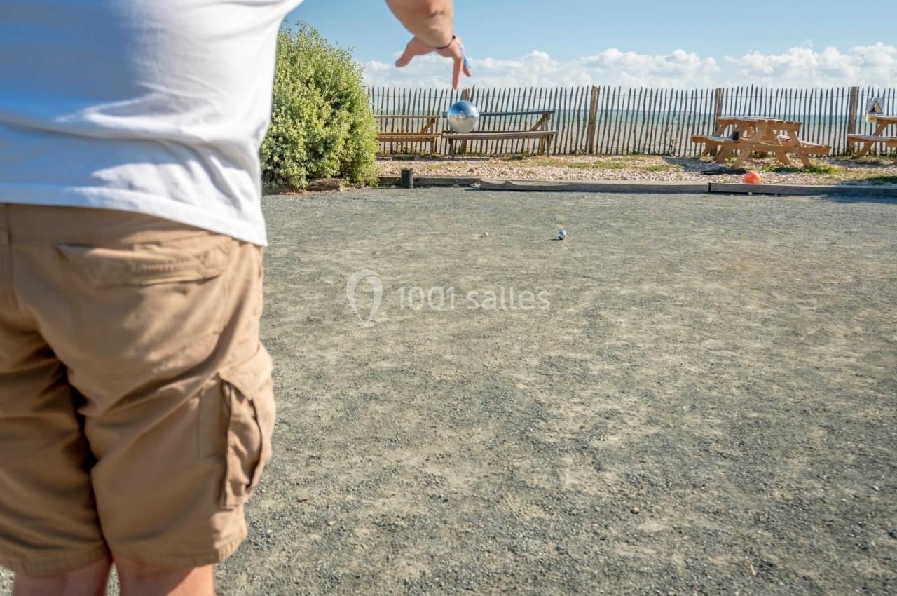 Un joueur de pétanque lance une boule sur un terrain en gravier, avec des tables de pique-nique en arrière-plan.